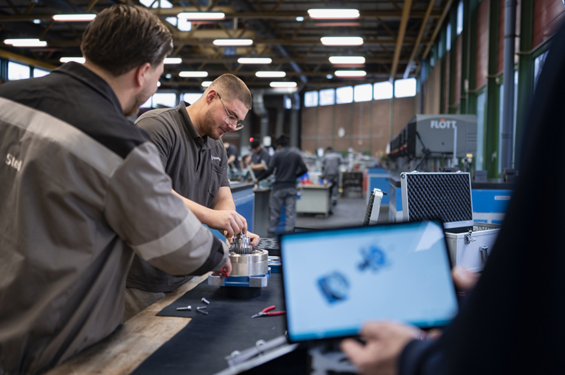 Ausbildung bei thyssenkrupp Steel: links stehen zwei Azubis in Kluft an der Werkbank, im Vordergrund ist unscharf ein Tablet-Display zu sehen