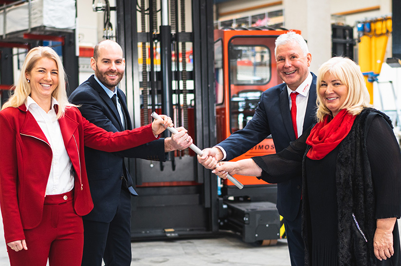 das Bild zweigt vier weiße Menschen: links stehen ein Mann und eine Frau mittleren Alters (die neuen Geschäftsführer von Dimos). Sie nehmen einen Staffelstab in die Hand, den sie von ihren Eltern (rechts im Bild) erhalten. Im Hintergrund ist ein Gabelstapler zu sehen.