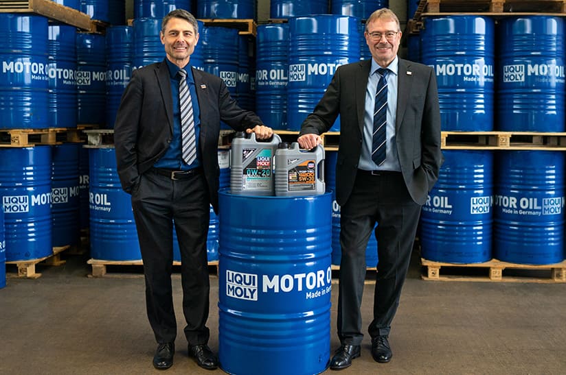 Zu sehen sind zwei weiße Männer im Anzug (die Geschäftsführer von Liqui Moly), die mit einer blauen Tonne und vor einer Wand aus blauen Tonnen posieren.