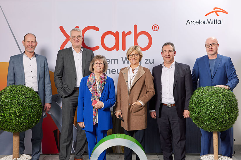 CO2-armer Stahl führt EWM und AcelorMittal zusammen. Im Bild ist Führungspersonal der beiden Unternehmen zu sehen, dazu im Vordergrund ein Stahlcoil und im Hintergrund ein Firmenlogo.