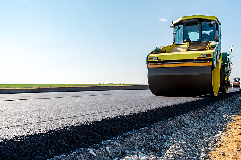 Das Bil zeigt am rechten Bildrand eine gelbe Walze, die eine frisch geteerte Straße walzt. Im Straßenbau werden Gesteinskörnungen aus Eisenhüttenschlacke seit Jahrzehnten im eingesetzt.
