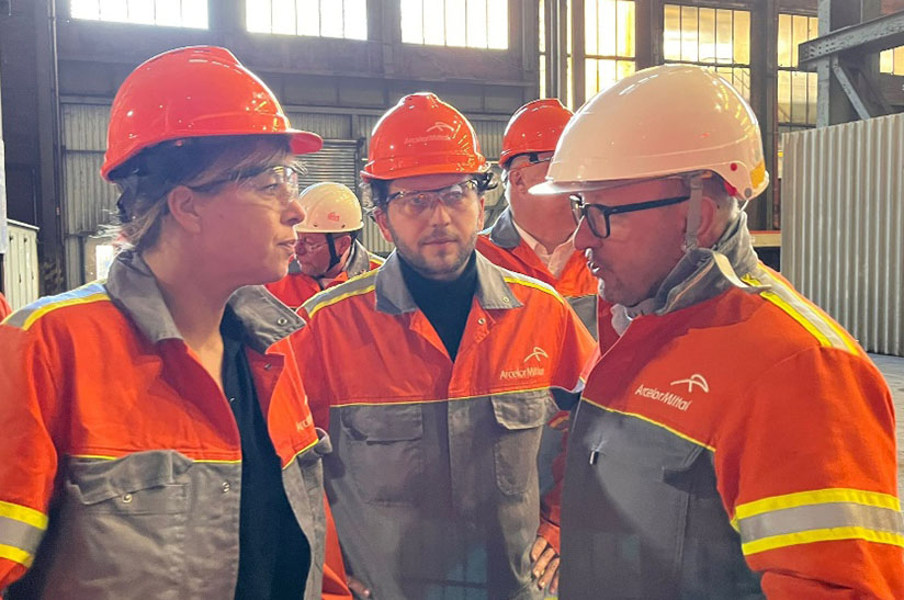 Das Bild zeigt im Vordergrund drei Menschen, die Helme tragen und Jacken des Stahlerzeugers ArcelorMittal: Mona Neubaur (links) und Felix Banaszak (Mitte) sowie Paul Tetteroo