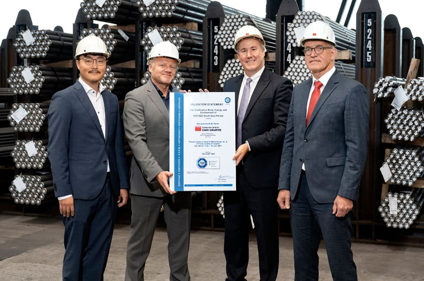 Das Bild zeigt vier männliche Anzugträger mit weißem Helmen, die in einem Lager mit Stahl stehen. Sie lächeln in die Kamera und die beiden in der Mitte halten gemeinsam eine große Urkunde. Es handelt sich hier um das Führungspersonal der GMH Gruppe und von TÜV Süd.