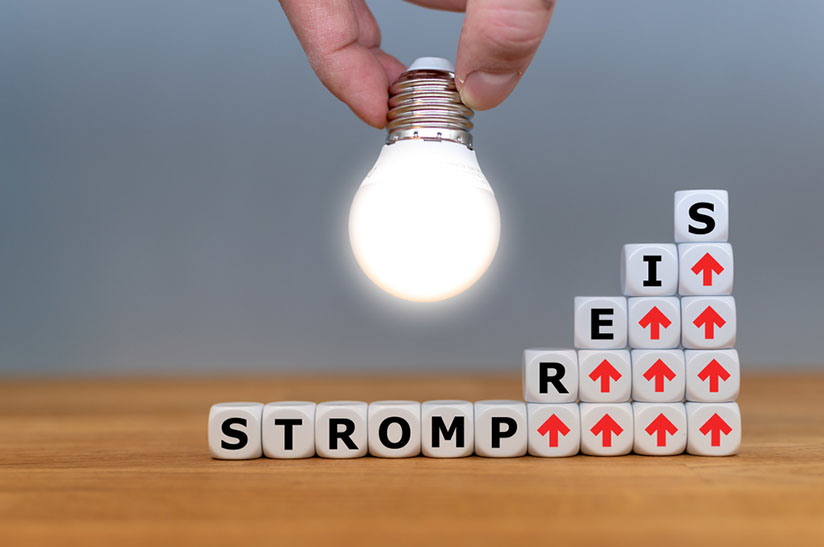Das Bild zeigt Würfel mit Buchstaben und roten Pfeilen, die für den steigenden Strompreis stehen. Eine helle Glühbirne soll die Idee der Siempelkamp Giesserei andeuten, wie das Problem gelöst werden kann.