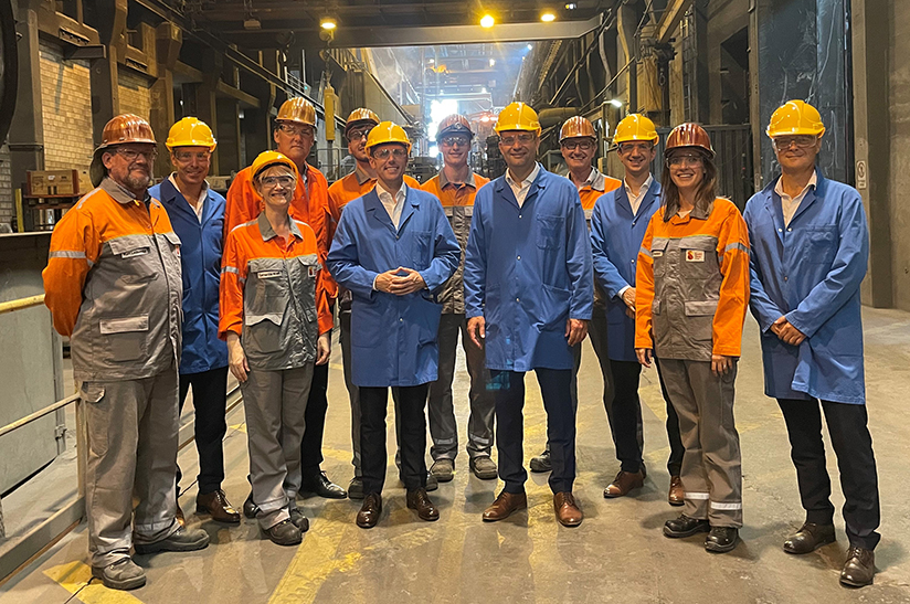 Das Bild zeigt eine Gruppe von Menschen im stahlwerk-szene-transformationsgipfel-aufruf der Swiss Steel Group. Mittig mit blauen Kitteln stehen links der US-Botschafter Scott Miller und der Luzerner Regierungspräsident Fabian Peter. Die Personen in orange-grauer Sicherheitskleidung gehören zum Unternehmen.