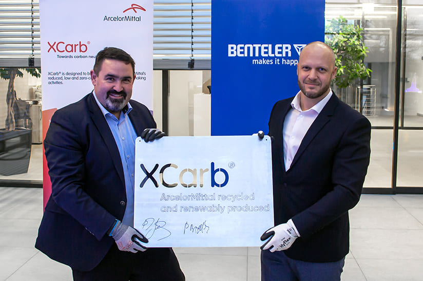 Das Bild zeigt zwei männliche, weiße Mitarbeiter der Unternehmen ArcelorMittal und Benteler, die gemeinsam ein Schild mit dem Schriftzug eines Produktnamens hochhalten