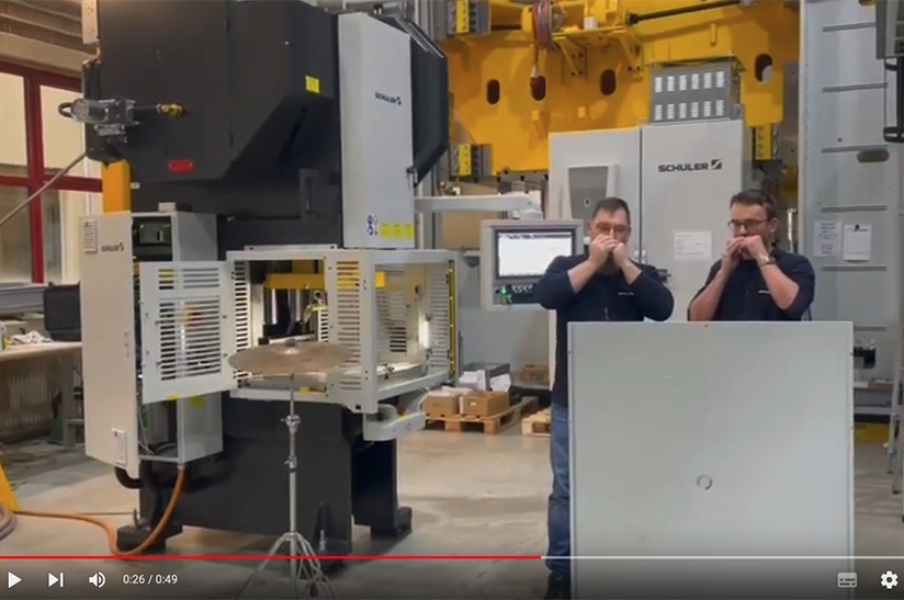 Das Bild zeigt einen Ausschnitt aus einem YouTube-Video der Schuler Group. Zwei Mitarbeiter spielen Mundharmonika und im Hintergrund hämmert eine Maschine auf ein Becken.