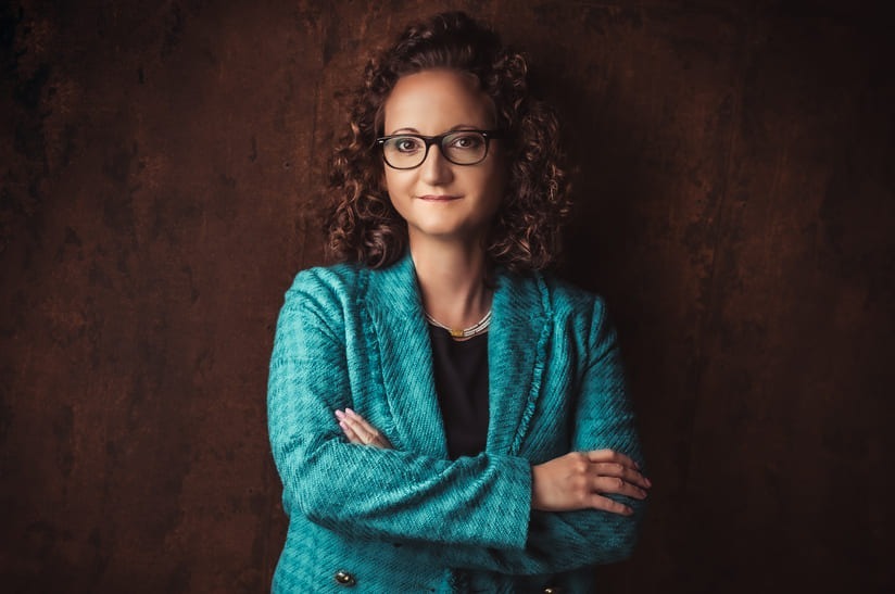 Das Bild zeigt eine Frau, Dr. Claudia Konrads, im Business-Outfit. Sie hat lockiges Haar und trägt eine Brille.