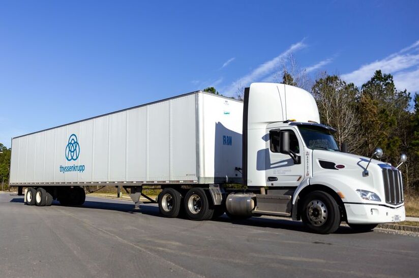 LKW mit Stahlcontainer von thyssenkrupp, blauer Himmel, Bäume im Hintergrund.