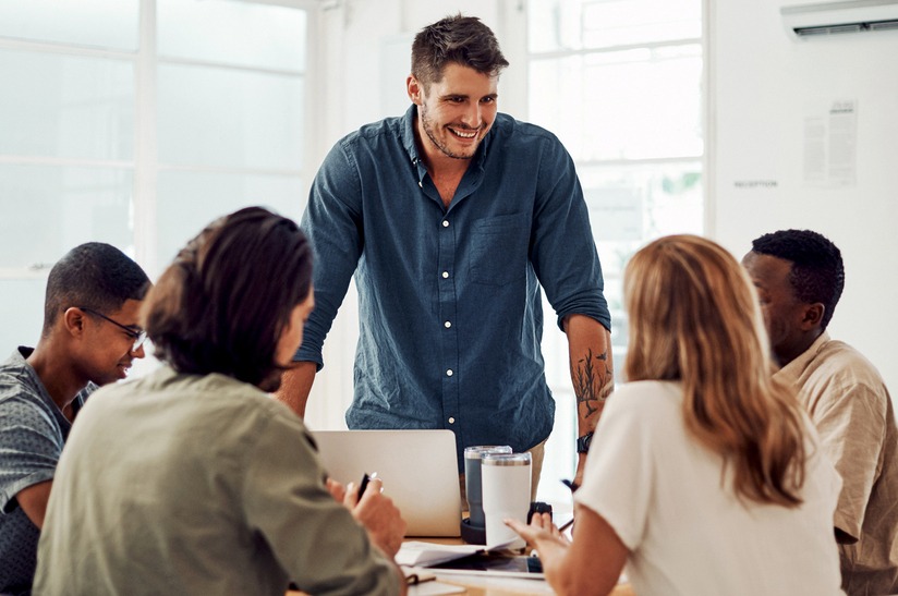 Das Foto zeigt fünf junge Erwachsene in einer Besprechung. Vier sitzen und hören zu, einer steht und spricht. Es geht um gewaltfreie Kommunikation, entsprechend lächelt der Sprecher.